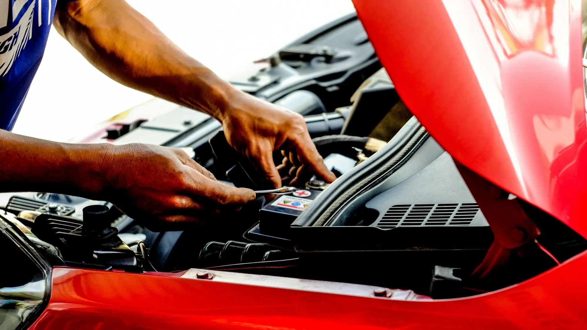 Mechanic's hands working under the raised hood of a red car outdoors